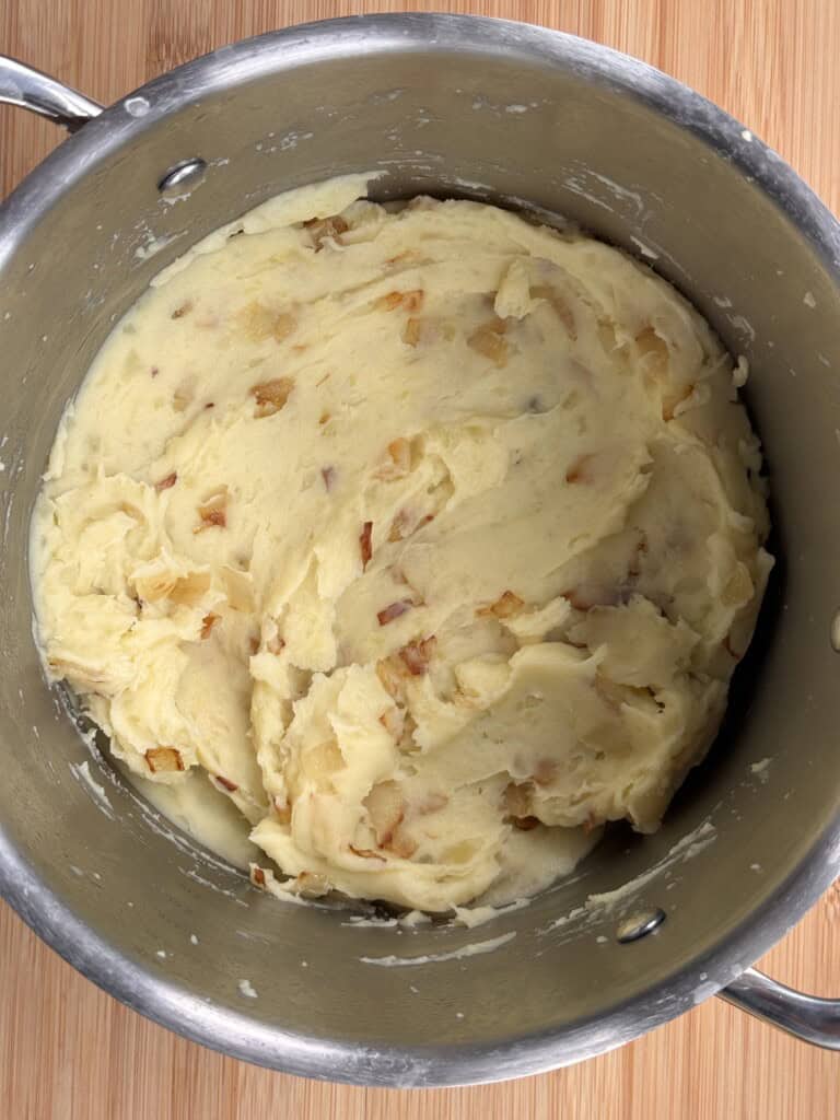A stainless steel saucepan filled with creamy mashed potatoes mixed with small pieces of cooked onions, sitting on a wooden surface.