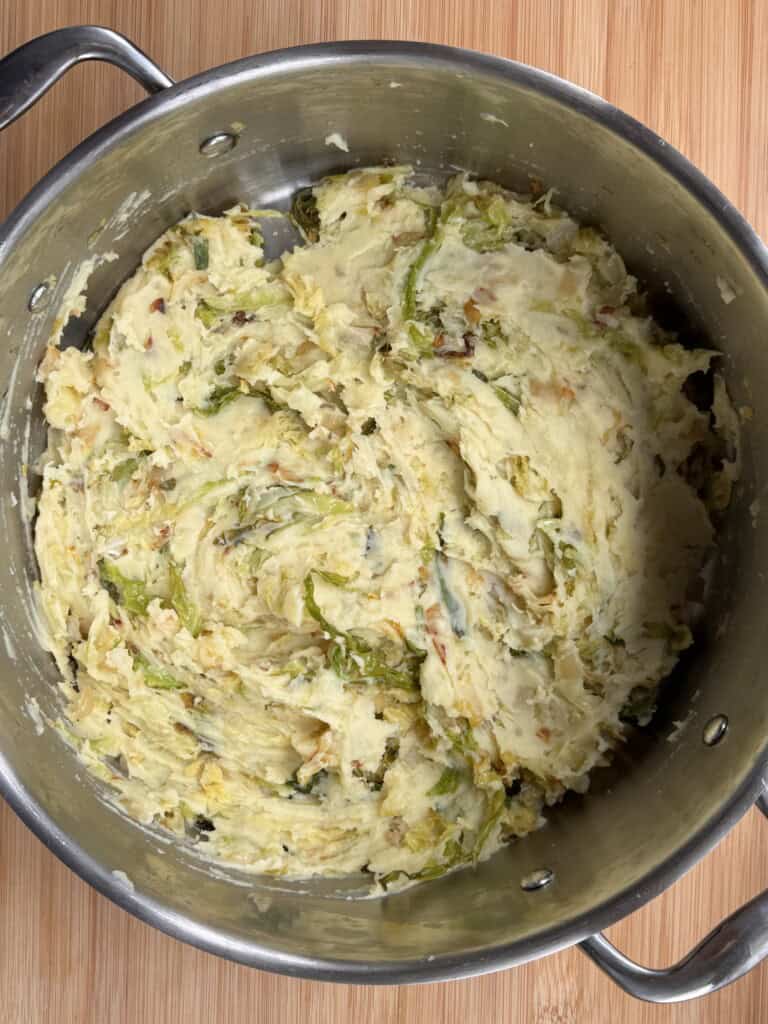 A large metal saucepan containing creamy mashed potatoes mixed with cooked leafy greens, sitting on a wooden surface.