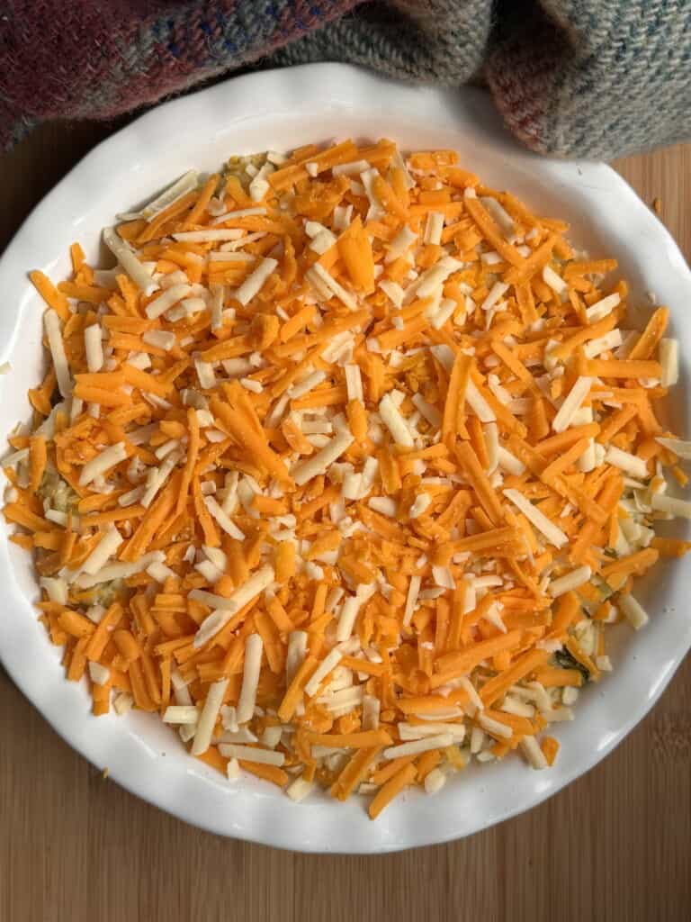 A white dish filled with a mixture topped with grated orange and white cheese on a wooden surface, with part of a colourful woven cloth visible at the top.