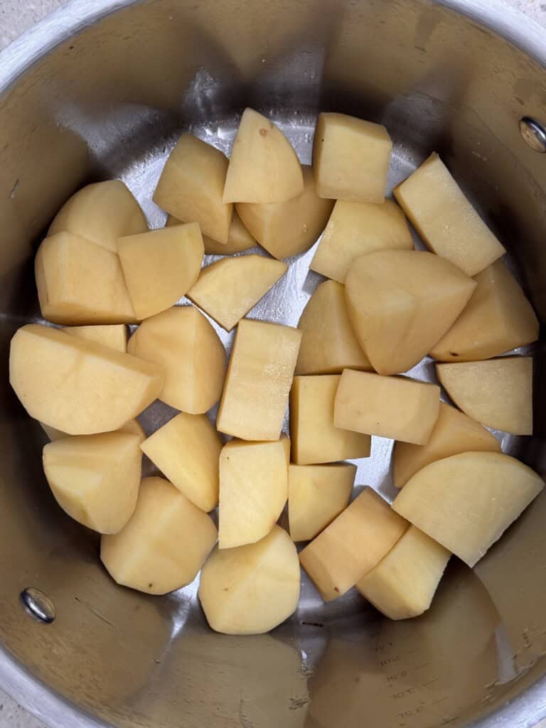 Chopped raw potatoes in large chunks are spread out inside a stainless steel saucepan, ready for cooking.