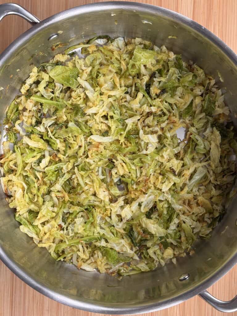 Shredded cabbage sautéed until lightly browned in a large stainless steel pan, sitting on a wooden surface.