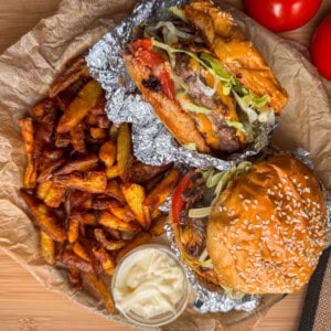 Two burgers with lettuce and tomato, one in a sesame bun and one in foil, served with a pile of crispy chips and a small pot of mayonnaise on greaseproof paper. Whole tomatoes are visible in the background.