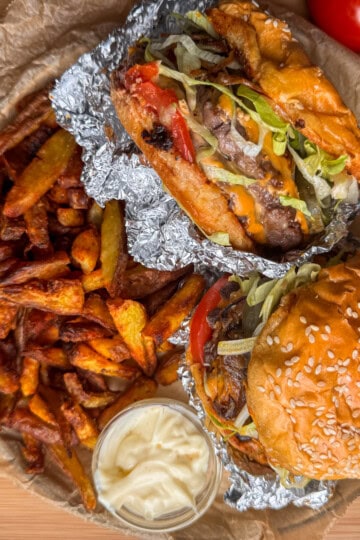 Two burgers with lettuce and tomato, one in a sesame bun and one in foil, served with a pile of crispy chips and a small pot of mayonnaise on greaseproof paper. Whole tomatoes are visible in the background.