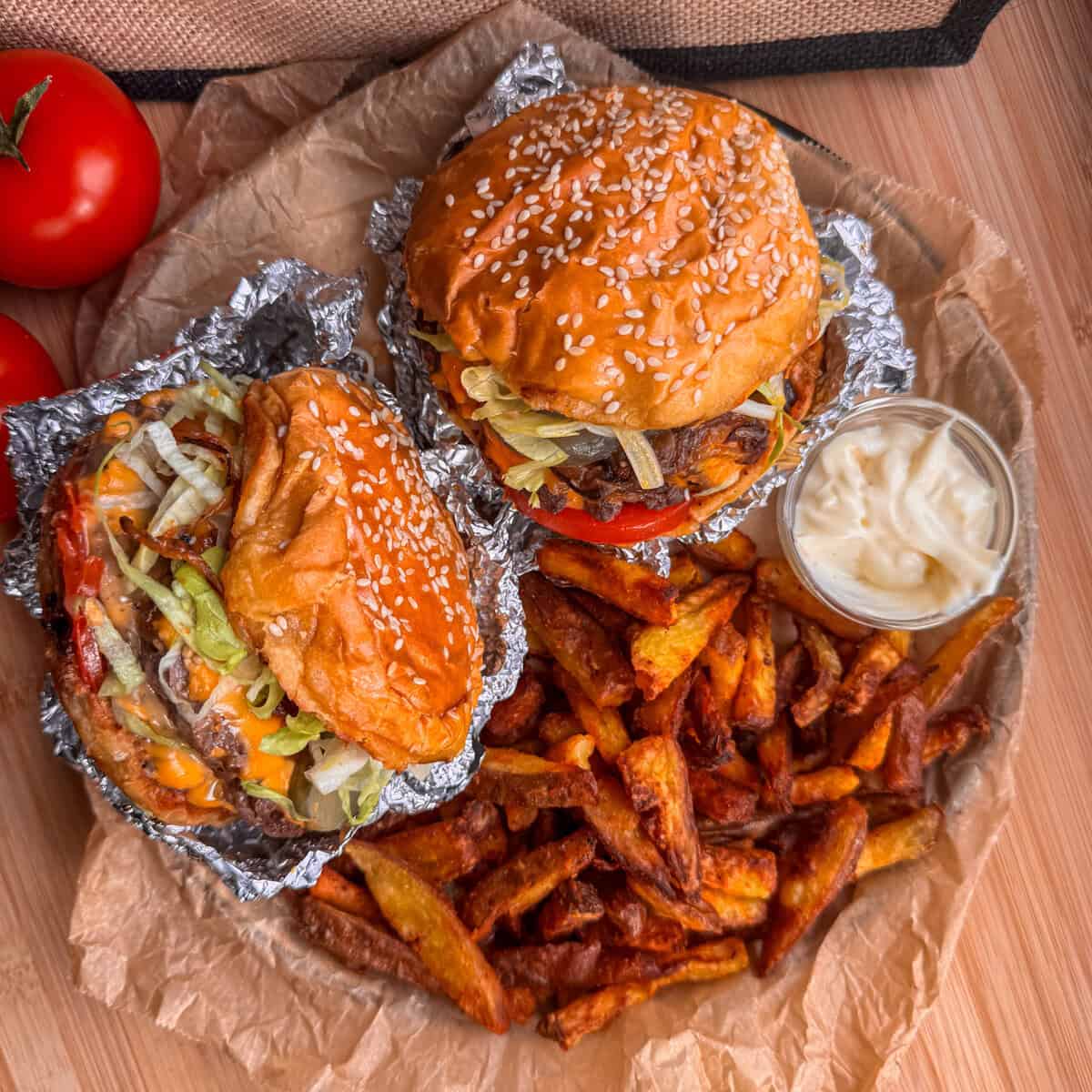 Two cheeseburgers with lettuce and tomato, wrapped partially in foil, served with a pile of chips and a side of mayonnaise on brown greaseproof paper, with two tomatoes in the background.