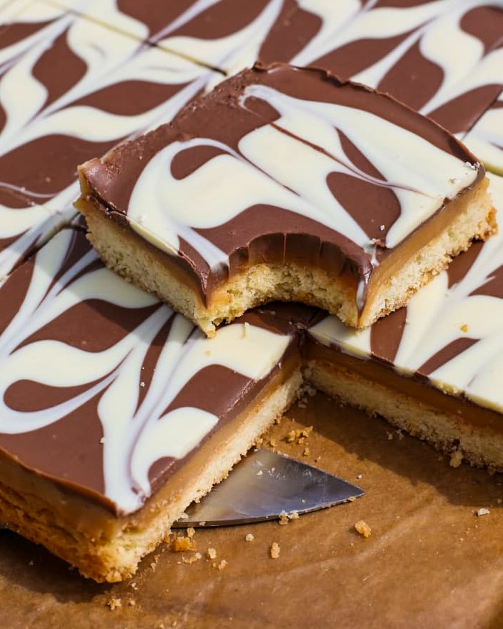 A close-up of chocolate caramel shortbread bars with a marbled dark and white chocolate topping. One square has a bite taken out, revealing layers of caramel and shortbread. A spatula is partially visible beneath the bars.