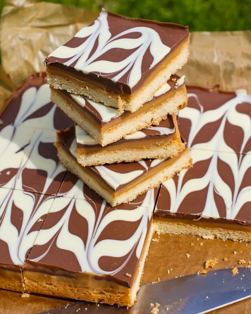 Stacked and sliced chocolate caramel shortbread bars topped with marbled milk and white chocolate swirl patterns, arranged on baking parchment with a grassy background.