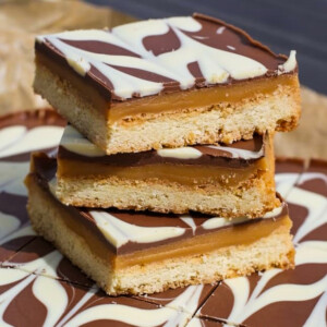 Three layered caramel shortbread bars stacked on top of each other, featuring a base, caramel centre, and chocolate topping with a white chocolate swirl pattern.
