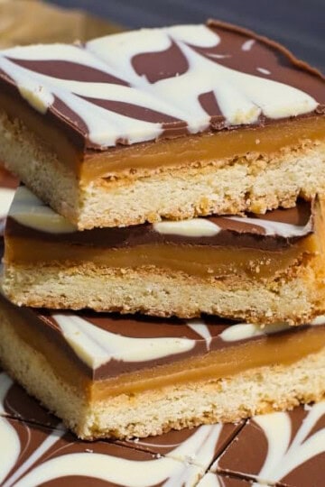 Three layered caramel shortbread bars stacked on top of each other, featuring a base, caramel centre, and chocolate topping with a white chocolate swirl pattern.