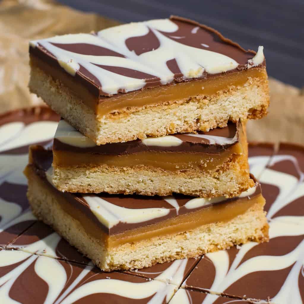 Three stacked caramel shortbread bars with a biscuit base, caramel layer, and chocolate topping decorated with a white chocolate swirl pattern. More bars are visible in the background.