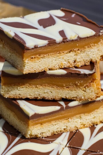 Three stacked caramel shortbread bars with a biscuit base, caramel layer, and chocolate topping decorated with a white chocolate swirl pattern. More bars are visible in the background.