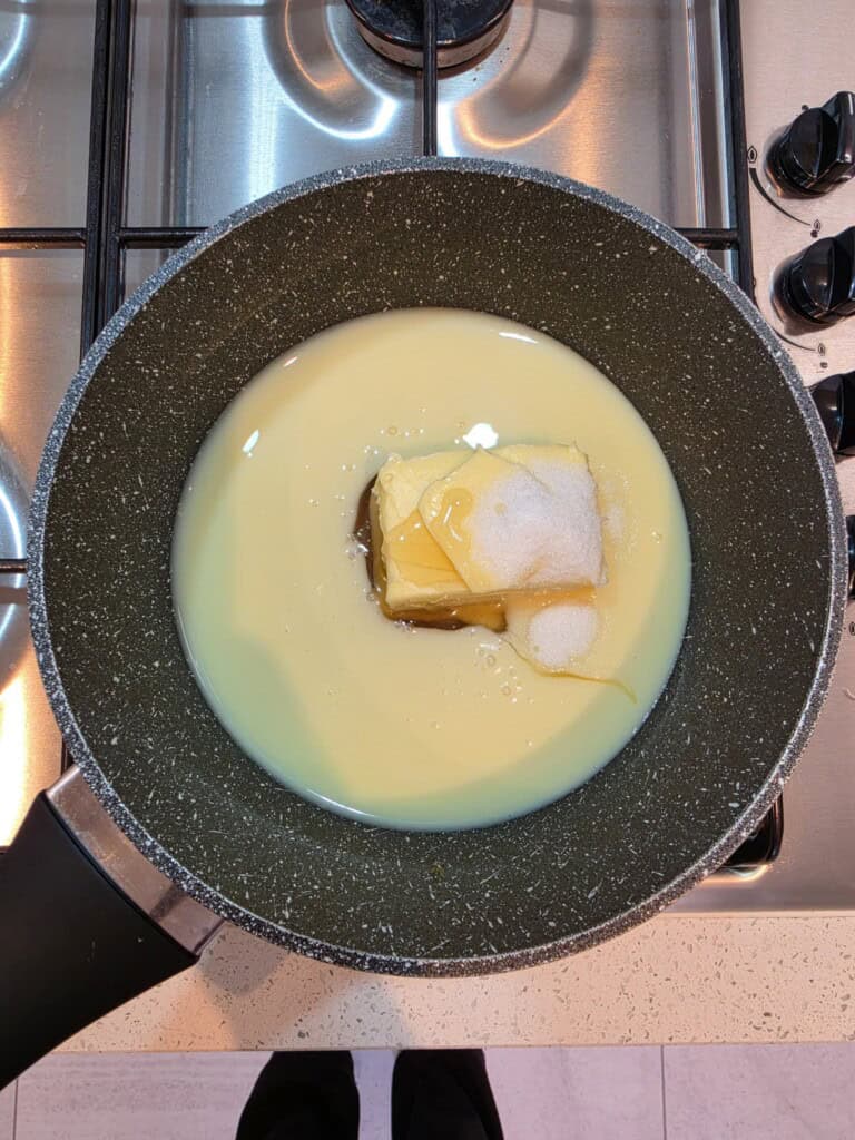 A non-stick saucepan on a hob contains sweetened condensed milk, a knob of butter, sugar, and honey before mixing or heating.