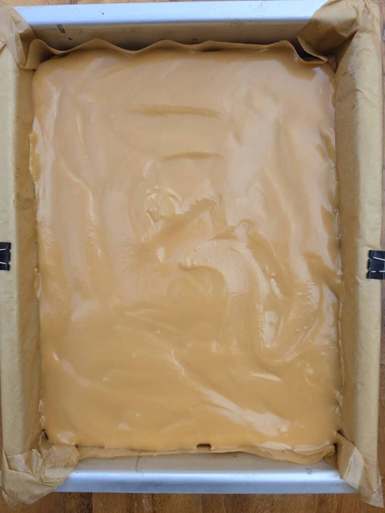 A square baking tin lined with greaseproof paper is filled with a smooth, light brown mixture, possibly peanut butter or caramel fudge, ready to be baked or set.