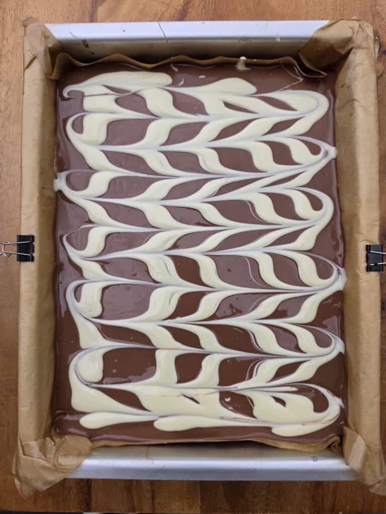 A rectangular tray of chocolate with a marbled pattern of white and milk chocolate swirled on top, lined with baking parchment and secured with bulldog clips on a wooden surface.