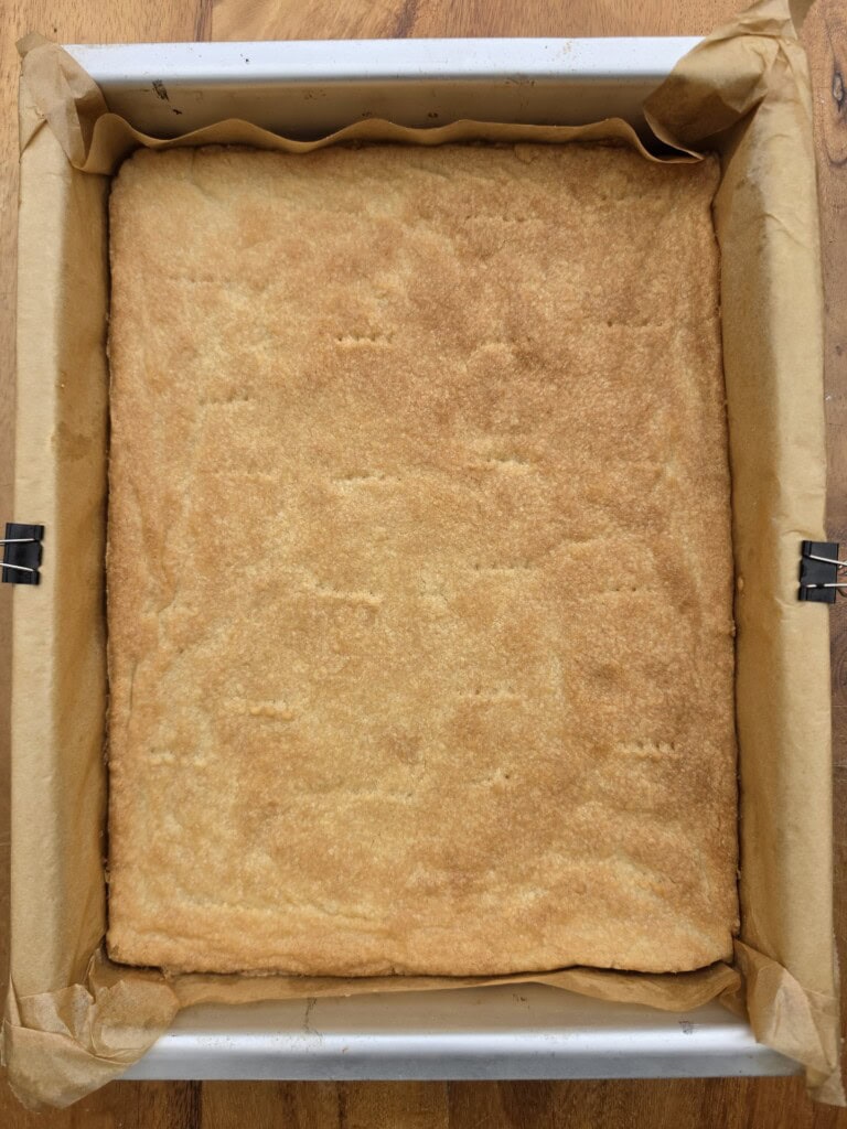 A rectangular tray lined with baking paper holds a large, evenly baked, golden brown shortbread slab with a slightly textured surface.