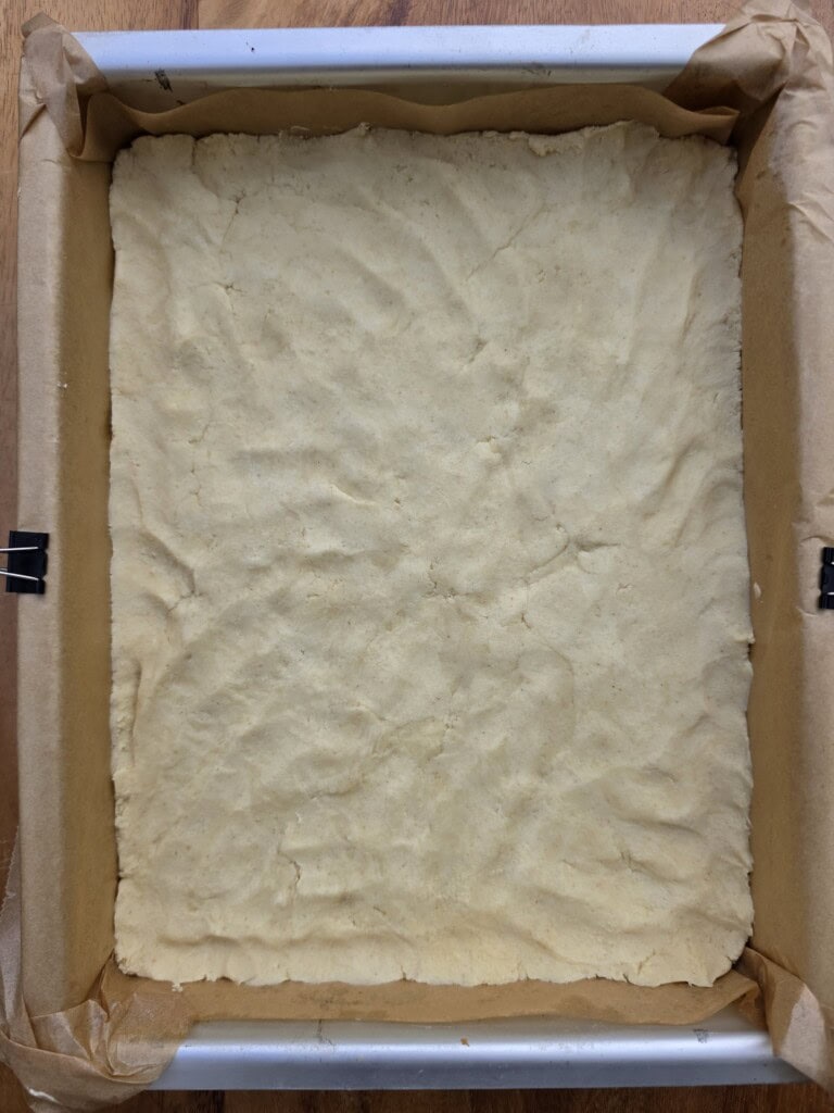 A baking tin lined with greaseproof paper holds an even layer of unbaked dough, pressed flat to cover the bottom. The greaseproof paper is secured with clips on the sides of the tin.
