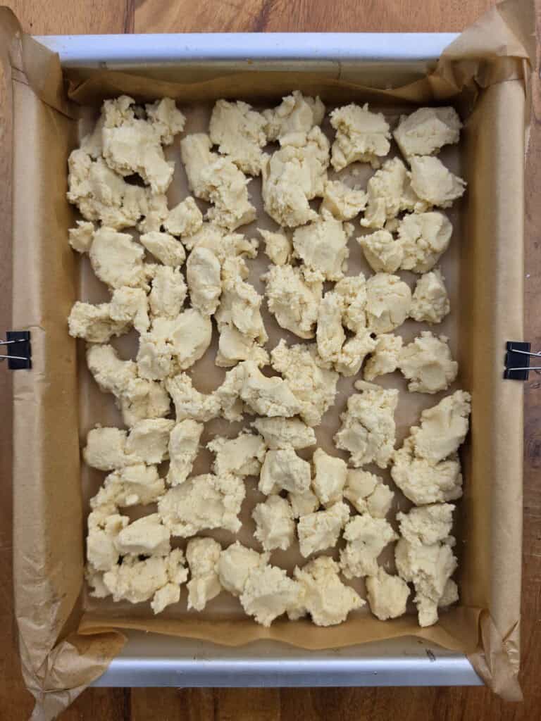 A baking tray lined with greaseproof paper holds uneven chunks of raw dough, spaced evenly apart and ready to be baked. The tray is secured with metal clips at the edges and sits on a wooden surface.