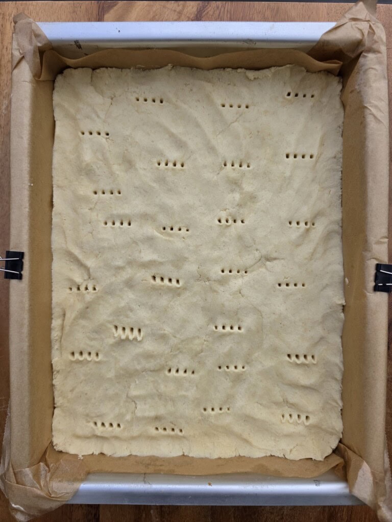 A rectangular baking tin lined with greaseproof paper holds a layer of raw shortbread dough, evenly pressed with small fork holes across the surface.