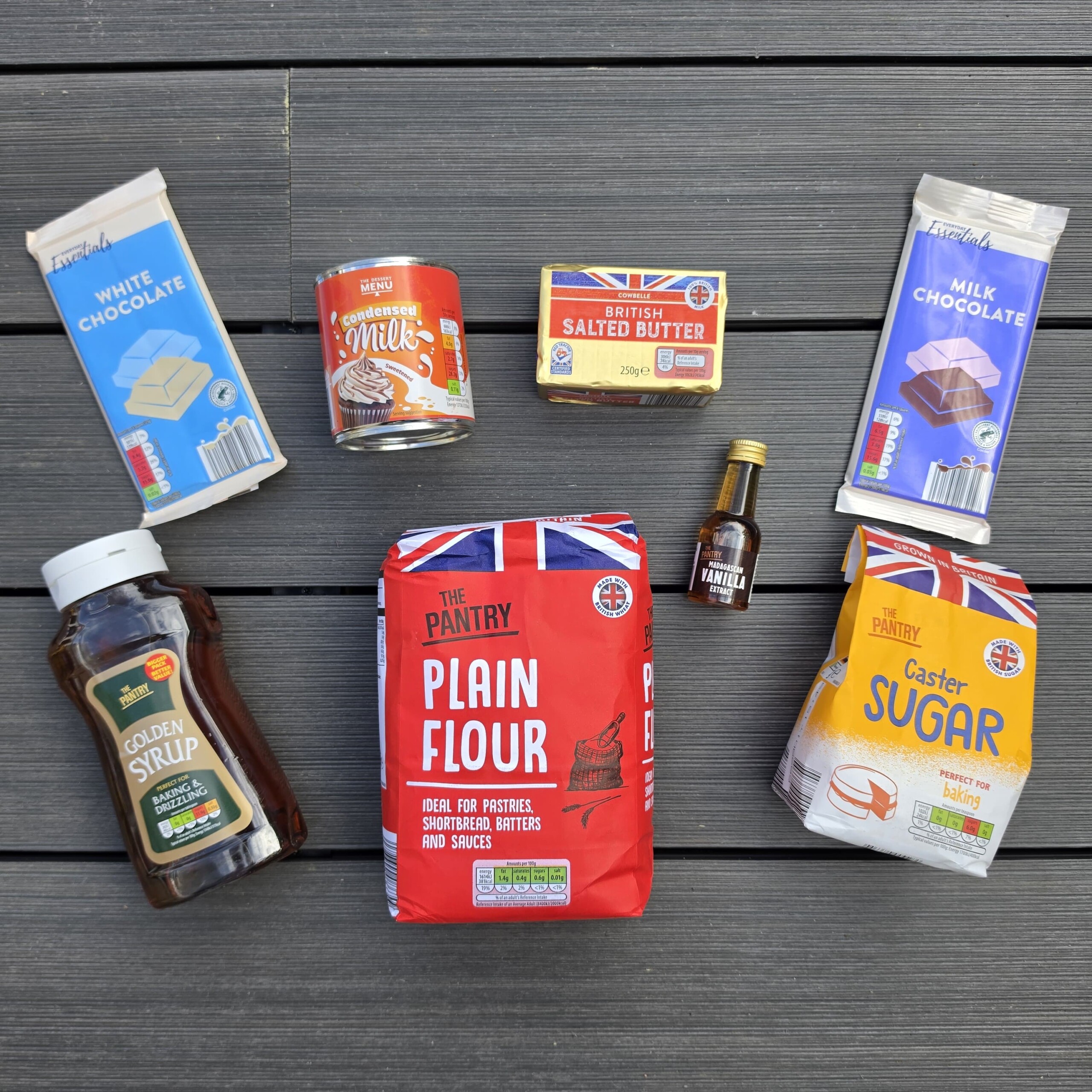 Baking ingredients arranged on a wooden surface, including plain flour, caster sugar, golden syrup, salted butter, vanilla extract, white and milk chocolate bars, and a tin of sweetened condensed milk.