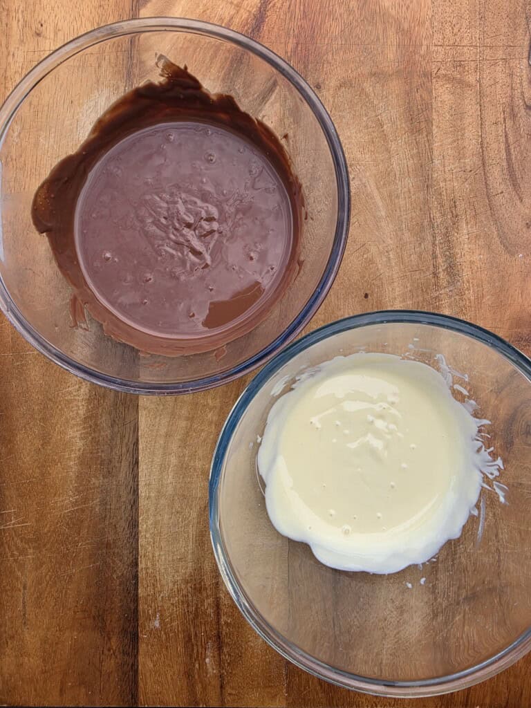 Two glass bowls on a wooden surface, one filled with melted milk chocolate and the other with melted white chocolate.