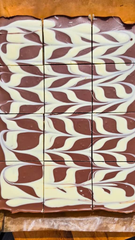 Chocolate squares topped with a marbled pattern of white and milk chocolate, divided into a 4 by 4 grid on greaseproof paper. The marbling creates a wave-like design across the surface.