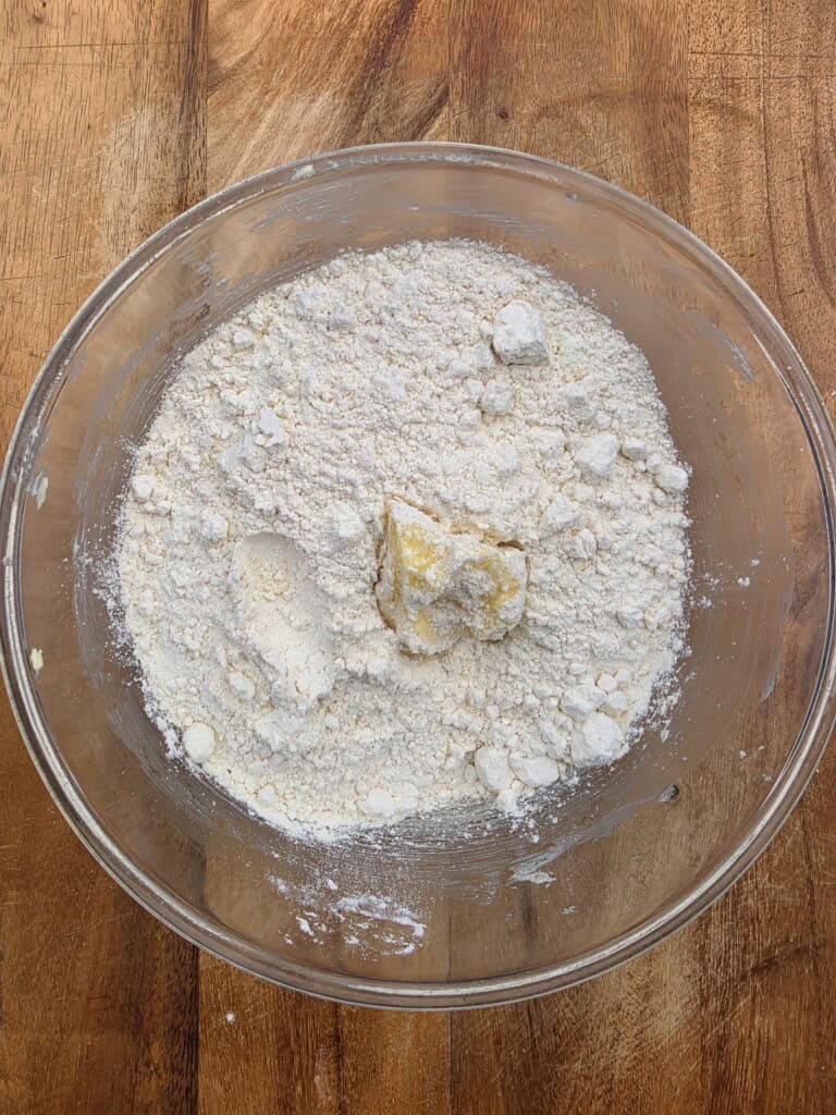 A glass bowl filled with flour and a chunk of butter sits on a wooden surface, showing the beginning stages of mixing baking ingredients.