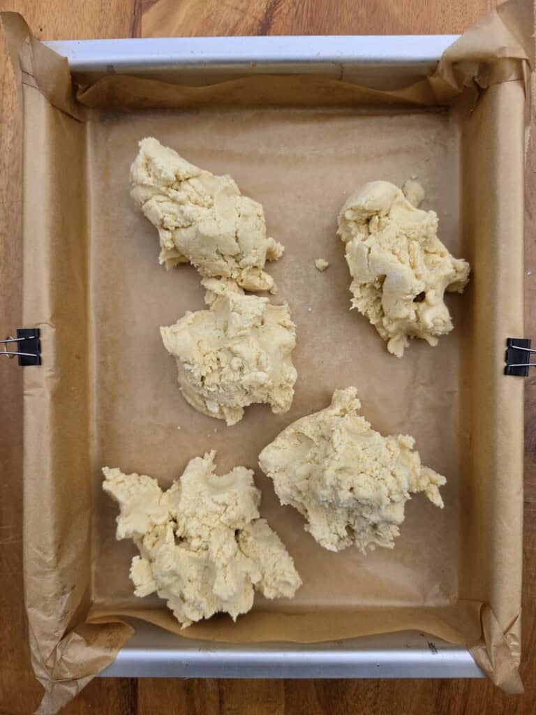 Five uneven scoops of pale, crumbly dough sit on a parchment-lined, square baking tray with metal binder clips securing the paper, all on a wooden surface.