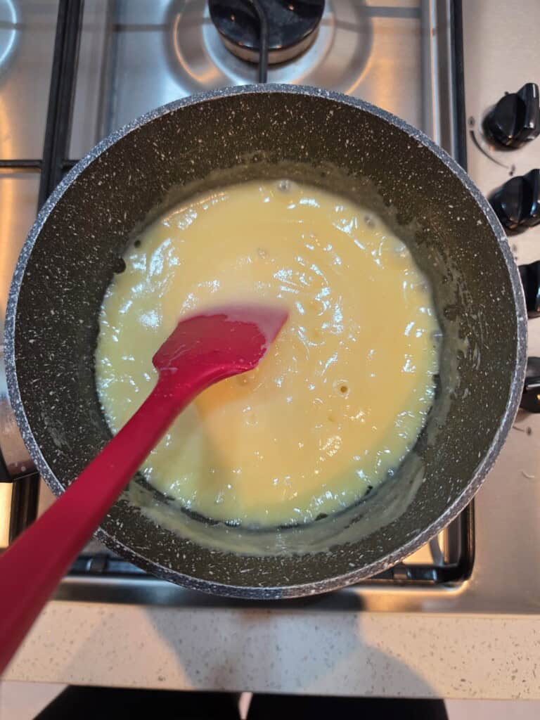 A red spatula stirs a thick, yellow mixture in a non-stick saucepan on a gas hob, possibly making a sauce or custard. The burner below is on, and cooker knobs are visible to the right.