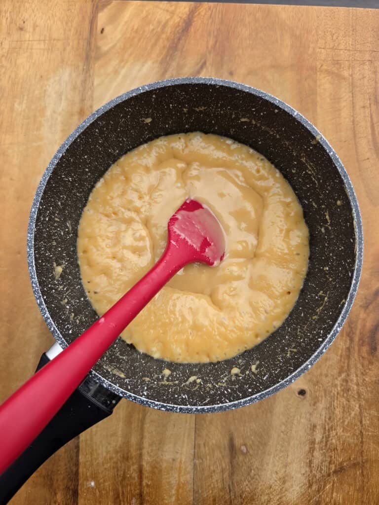 A red spatula stirs a yellow, thick liquid in a non-stick saucepan set on a wooden surface.
