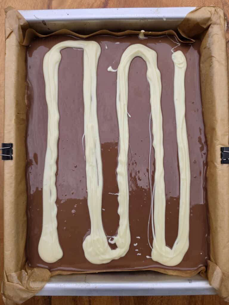 A baking tray lined with greaseproof paper holds a layer of smooth milk chocolate with three thick zigzag lines of white chocolate drizzled on top.