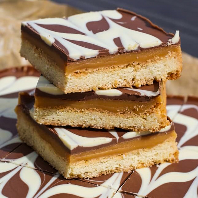 Three layered caramel shortbread bars stacked on top of each other, featuring a base, caramel centre, and chocolate topping with a white chocolate swirl pattern.