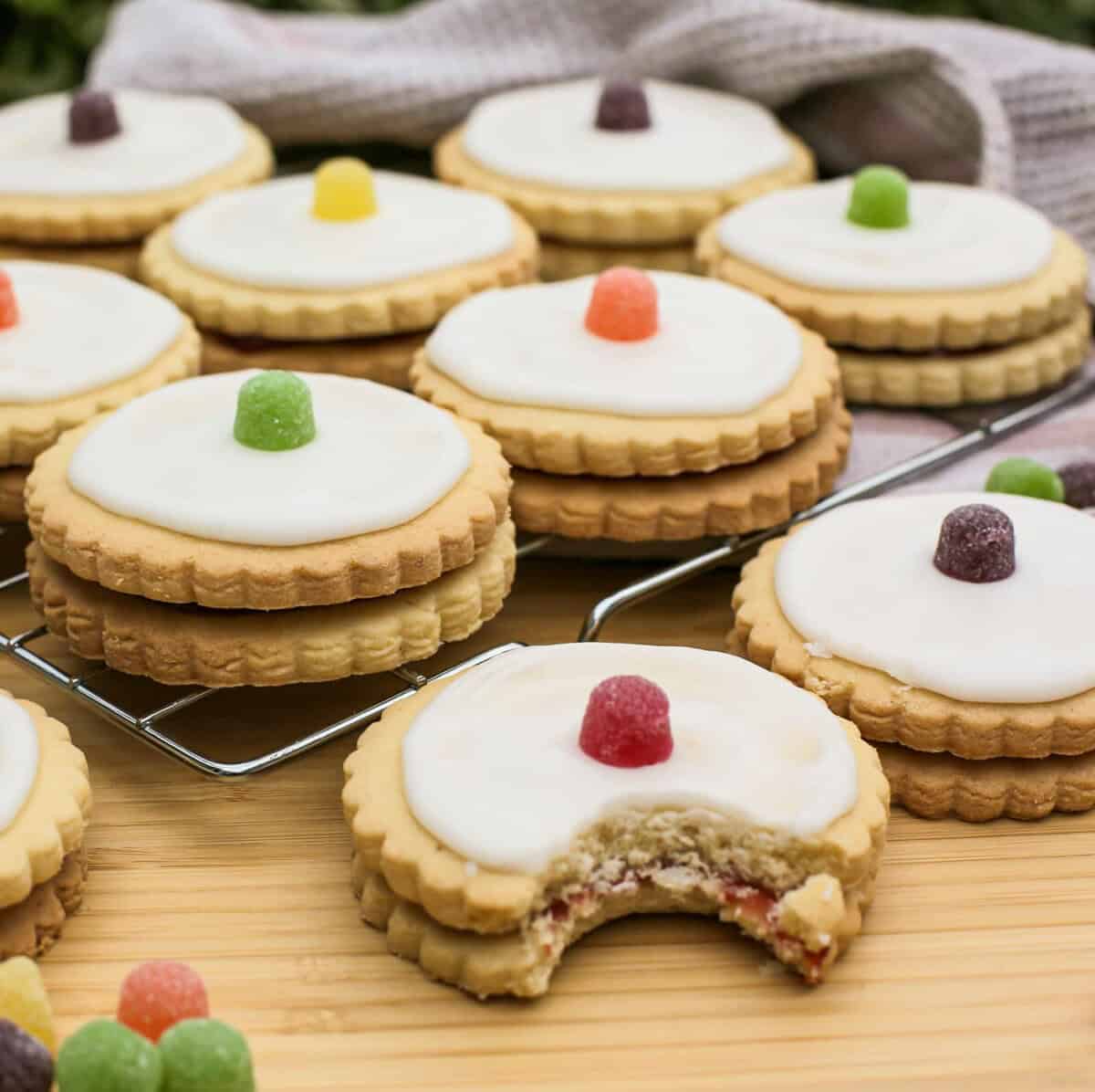 A group of round empire biscuits with scalloped edges and white icing, each topped with a colourful jelly sweet. One biscuit with a bite taken out sits in front, revealing a jam filling. The biscuits rest on a cooling rack and wooden surface.