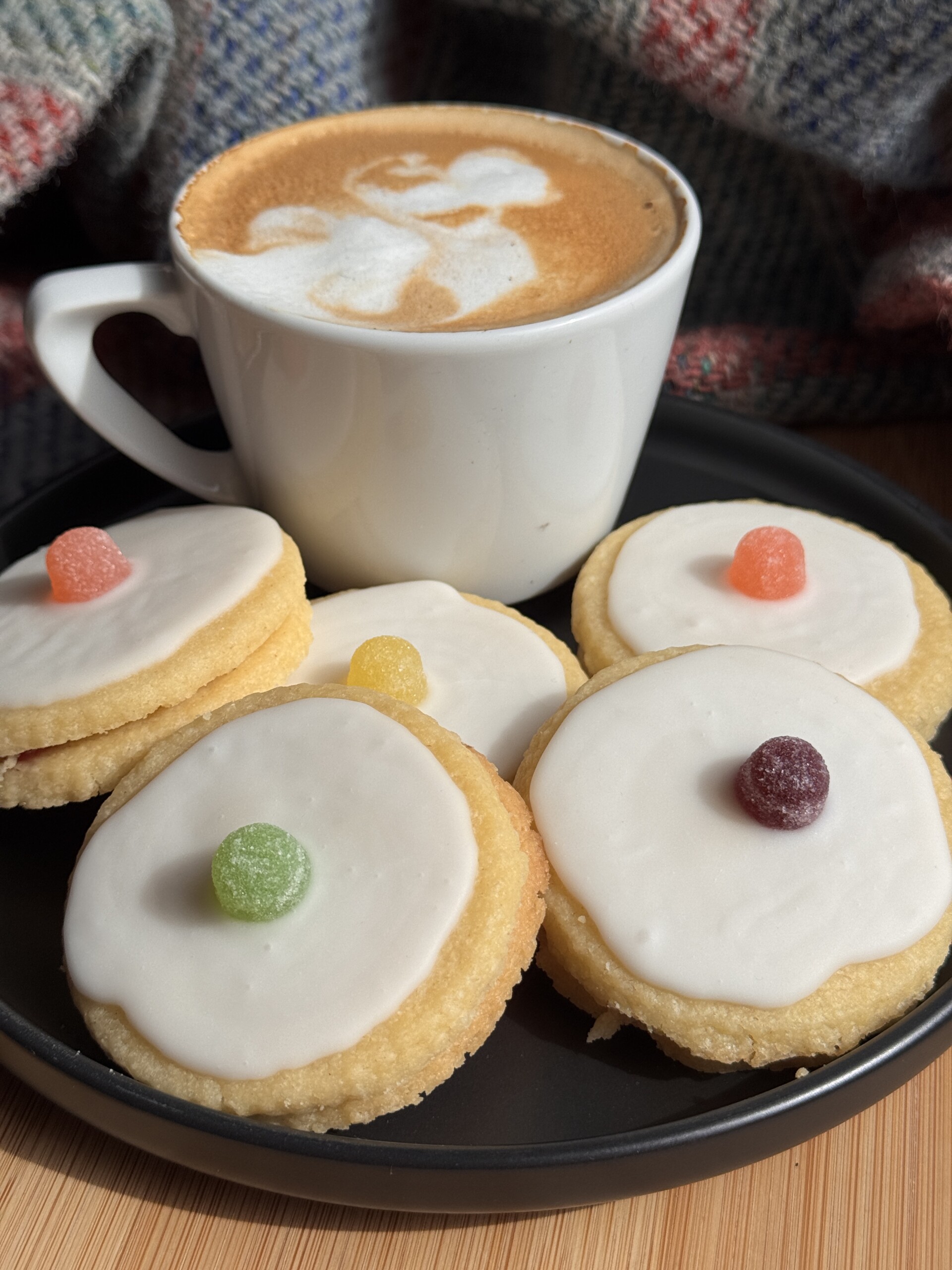 A white cup of cappuccino with foam art beside five empire biscuits topped with white icing and colourful gumdrops, all on a black plate with a chequered fabric in the background.