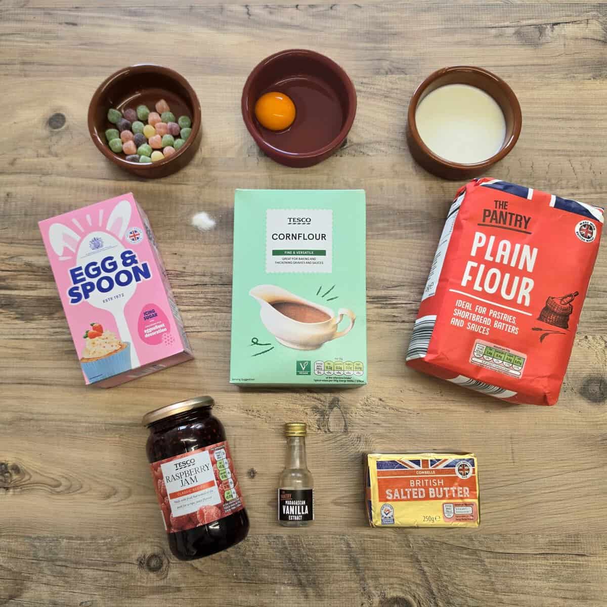 Various baking ingredients are arranged on a wooden surface, ready for making empire biscuits, including eggs, milk, butter, flour, cornflour, raspberry jam, vanilla essence, a box of cake mix, and a small bowl of colourful sweets.