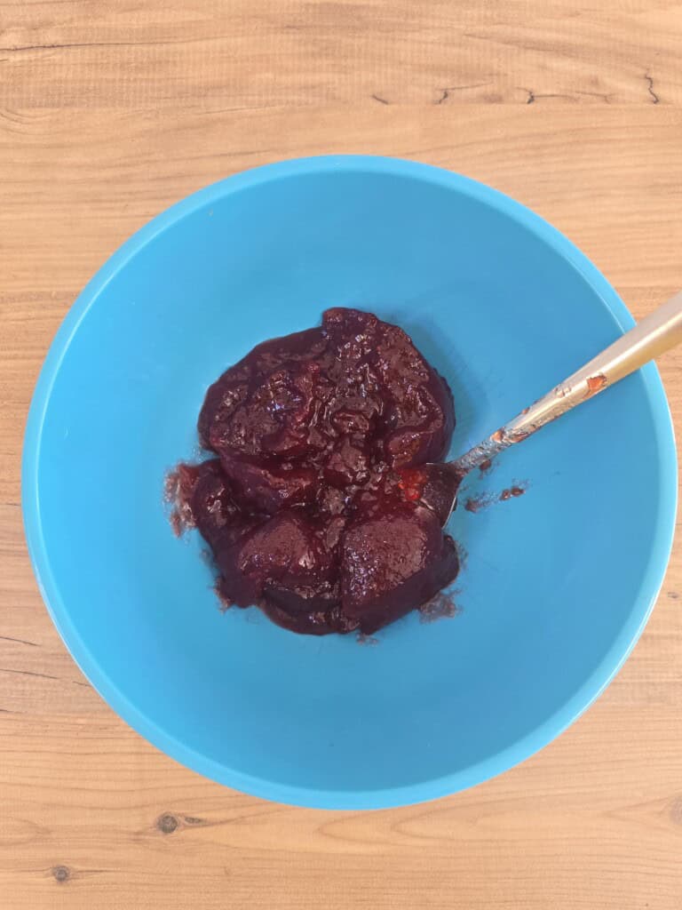 A blue bowl containing a thick, dark red chunky mixture with a metal spoon inside, placed on a wooden surface.