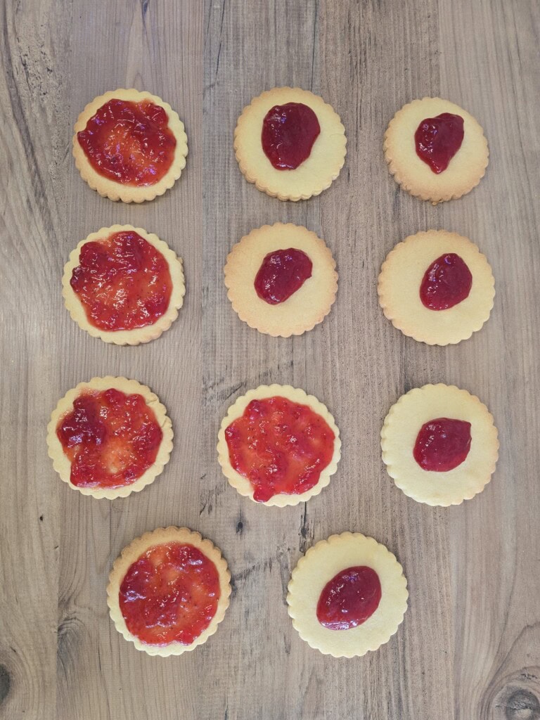 Twelve round biscuits are arranged in a grid on a wooden surface. The left column has biscuits with spread strawberry jam, the right column has plain biscuits with jam in the centre, and the middle column has plain biscuits with jam dollops.
