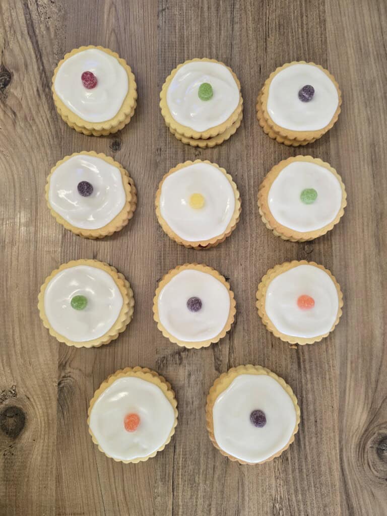 Twelve round iced biscuits, each topped with a single colourful jelly sweet, are arranged in a neat grid on a wooden surface.