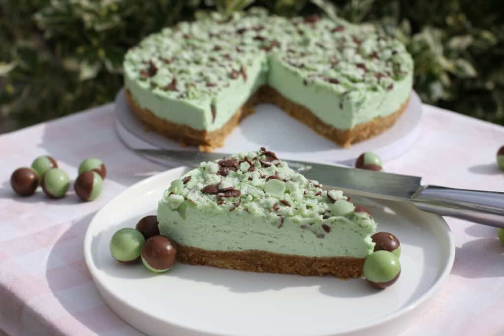 A slice of mint Aero cheesecake with a biscuit base sits on a plate, garnished with crushed chocolate and mint sweets, with a knife and the remaining cheesecake in the background.