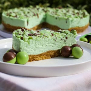 A slice of mint Aero cheesecake with a crumbly biscuit base, topped with chocolate and mint pieces, is served on a white plate with green and chocolate-coated sweets. The whole cake is in the background.
