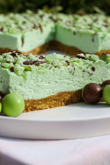 A slice of mint Aero cheesecake with a crumbly biscuit base, topped with chocolate and mint pieces, is served on a white plate with green and chocolate-coated sweets. The whole cake is in the background.