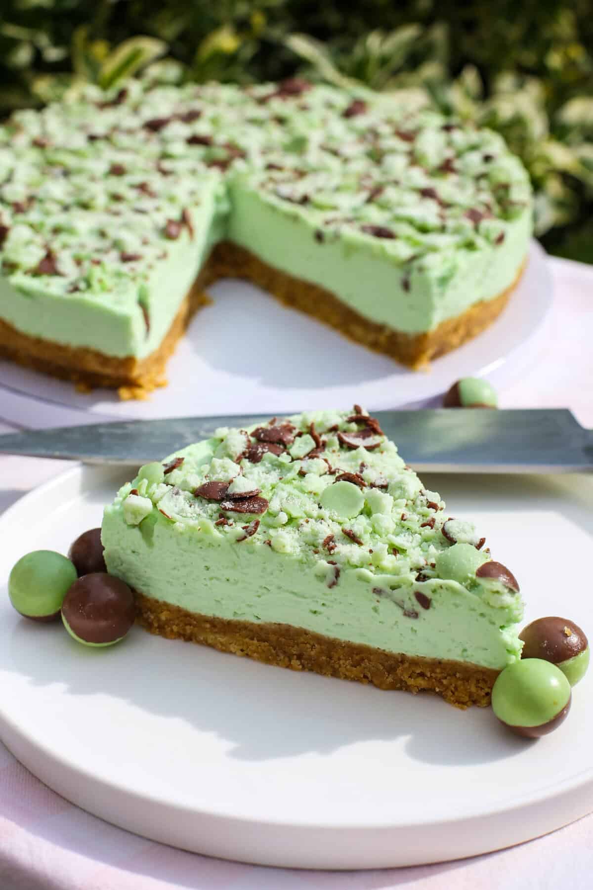 A slice of mint green mint Aero cheesecake with a crumbly biscuit base, topped with crushed chocolate and mint sweets, sits on a plate. The whole cheesecake and some round mint chocolates are visible in the background.