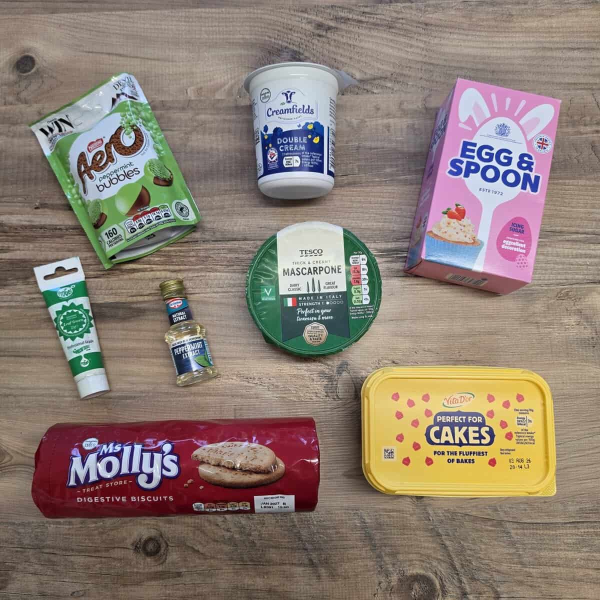 An assortment of baking ingredients on a wooden surface, including mascarpone, double cream, egg & spoon chocolate, Aero bubbles, green food colouring, vanilla, digestives, and Perfect for Cakes margarine.