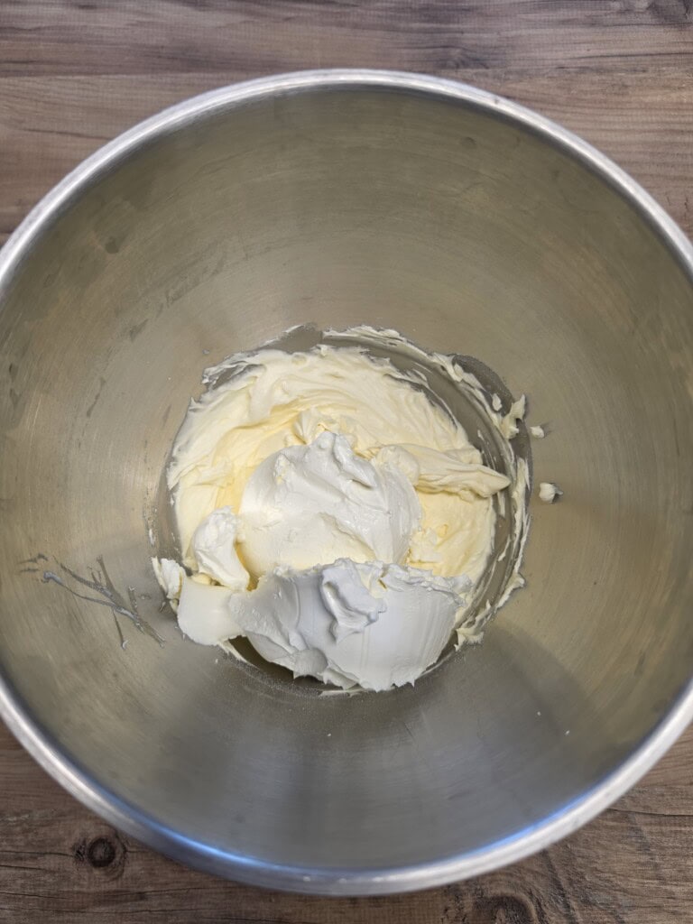 A metal mixing bowl containing a mound of softened cream cheese and butter, set on a wooden surface.