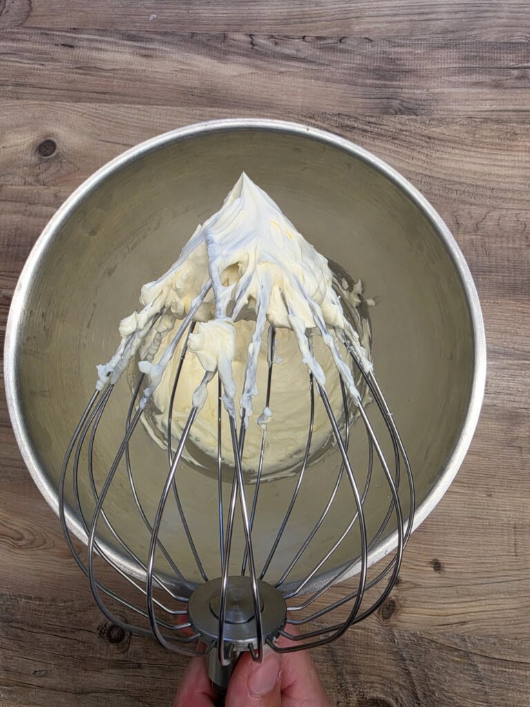 A hand holding a whisk with stiff whipped cream peaks above a metal mixing bowl on a wooden surface.