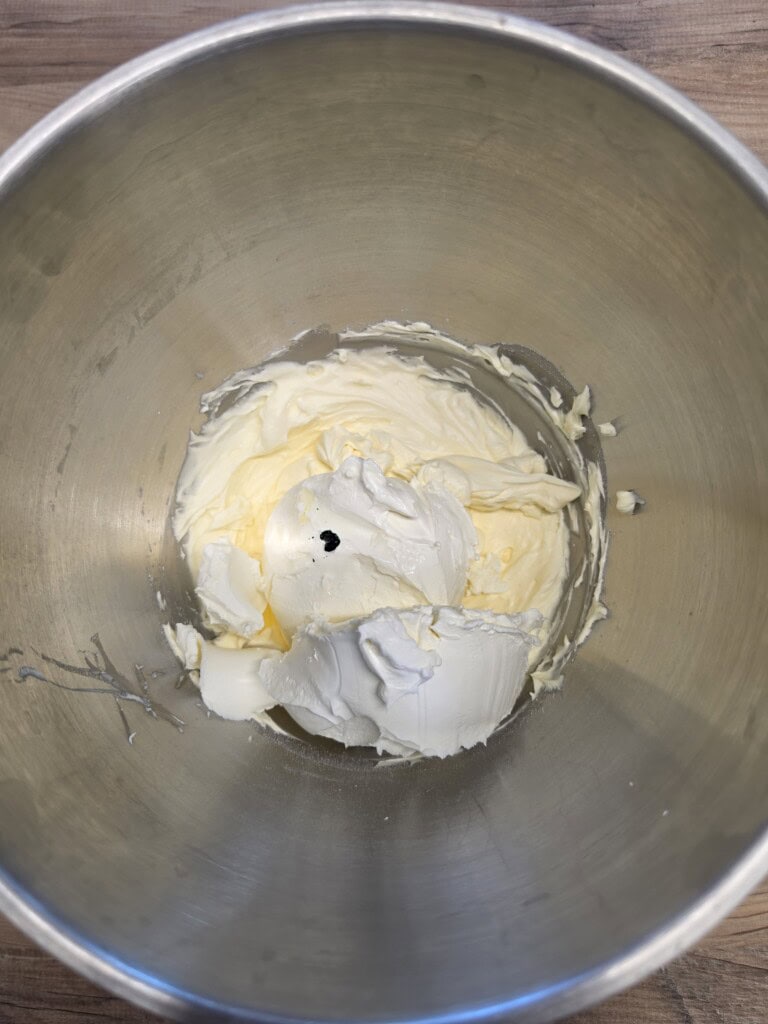 A metal mixing bowl containing mounds of cream cheese and butter with a small drop of black food colouring on top, placed on a wooden surface.