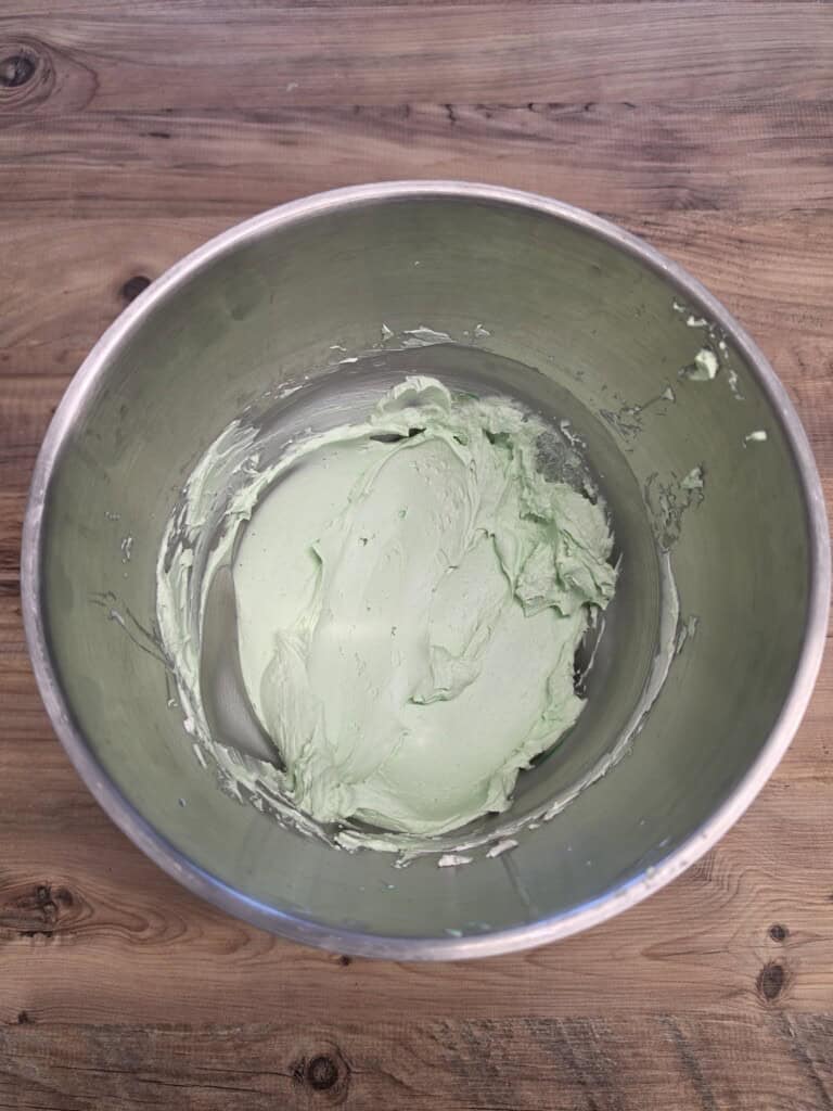 A mixing bowl containing pale green whipped icing sits on a wooden surface. The icing is smooth and partially spread around the sides of the bowl.
