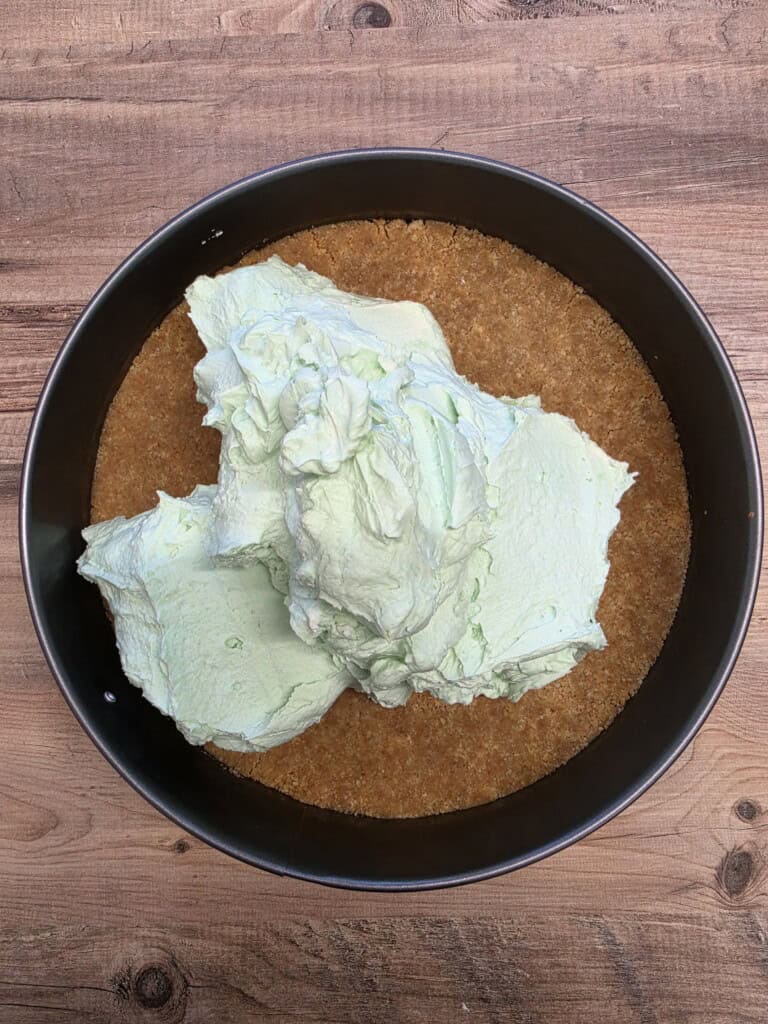 A round tin with a crumbly brown base on the bottom, topped with a large dollop of light green creamy filling, all placed on a wooden surface.