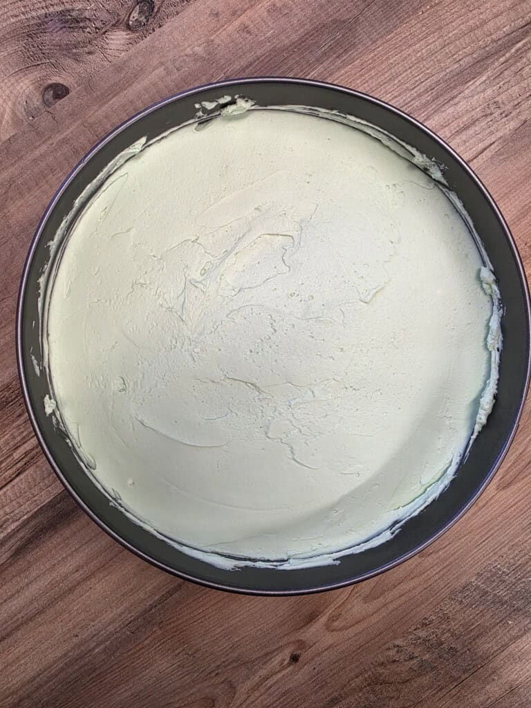A round tin filled with a smooth, pale green cheesecake sits on a wooden surface. The top of the cheesecake is even and creamy with slight texture marks.
