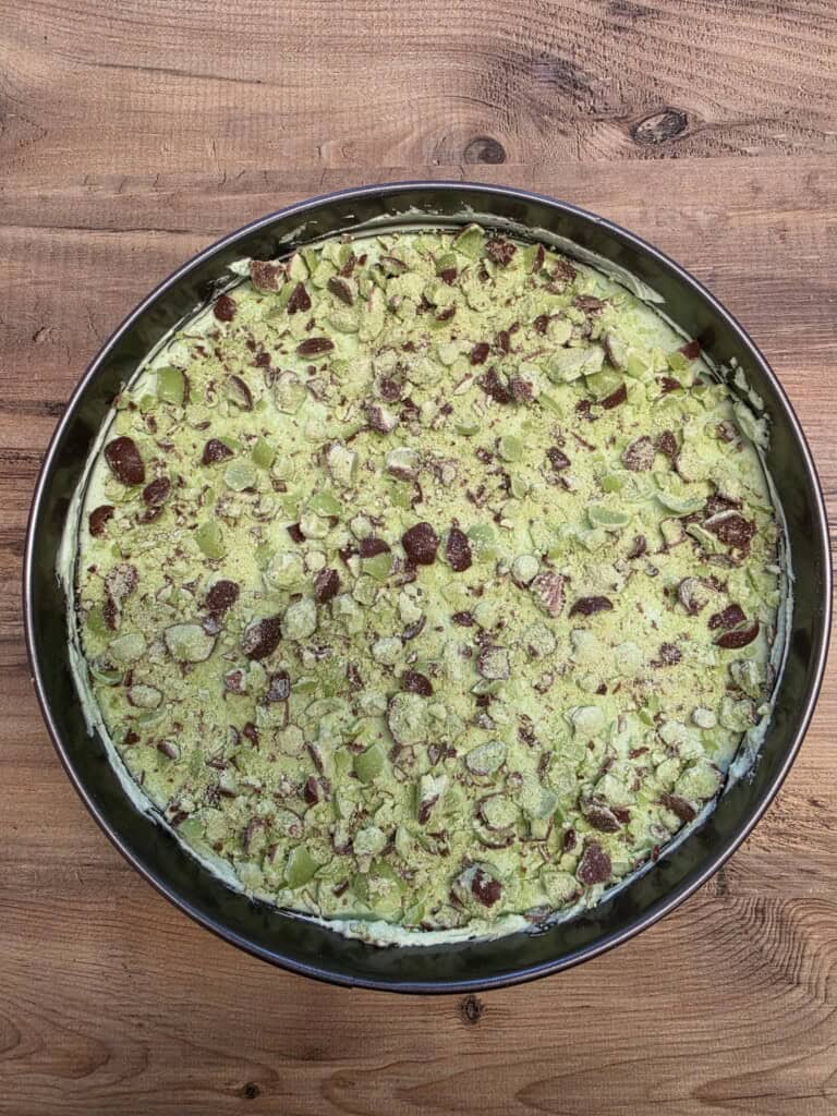 A round tin filled with a green mint Aero cheesecake topped with crushed mint aero balls, sits on a wooden surface. The cheesecake appears creamy and is evenly covered with the topping.