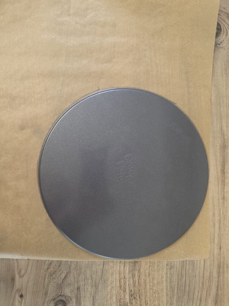 A round, dark grey baking tin is placed on a sheet of greaseproof paper on a wooden surface. The greaseproof paper is partially under the tin and extends to the left.