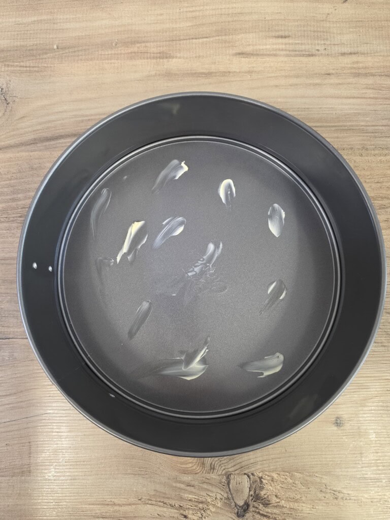 A round metal baking tin with a few smears of butter on the bottom, placed on a light wooden surface.